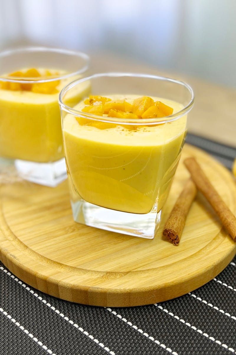 Home Two glasses of creamy mango lassi garnished with diced mango and cinnamon sticks.